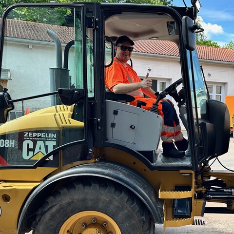 Mitarbeiter in orangefarbener Warnkleidung sitzt in einem Radlader und zeigt lächelnd den Daumen hoch