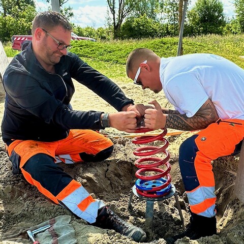 Zwei Mitarbeiter in Arbeitskleidung montieren gemeinsam eine rote Federwippe auf einem Spielplatz
