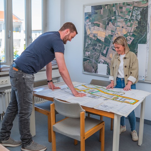Bauplanung – Zwei Mitarbeitende betrachten gemeinsam einen großen Stadtplan auf einem Tisch