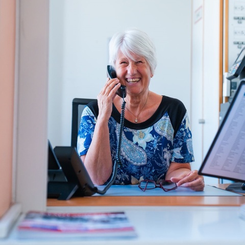 Ältere Dame mit Telefonhörer lächelt am Empfangsschalter