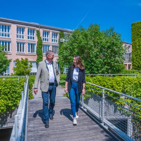 Brücke zwischen Gebäuden – Zwei Mitarbeitende gehen über eine Brücke zwischen begrünten Verwaltungsgebäuden