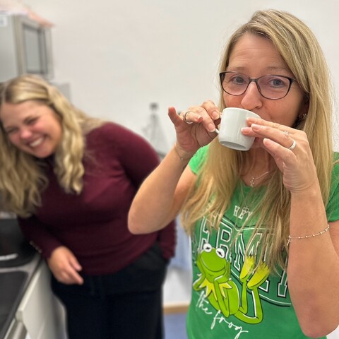 Kaffeepause – Zwei Mitarbeiterinnen in der Küche, eine trinkt Kaffee, die andere lacht im Hintergrund