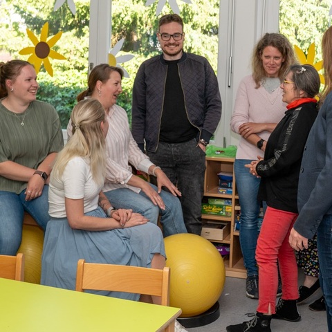Gruppe von acht Personen steht und sitzt in einem hellen Raum, lacht und unterhält sich