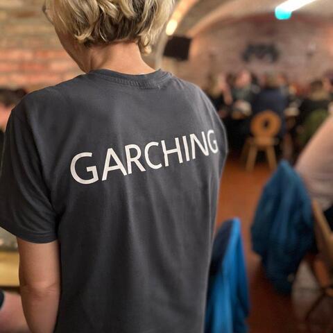 Rückansicht T-Shirt – Person mit grauem T-Shirt mit der Aufschrift „Garching“ in einem Veranstaltungsraum