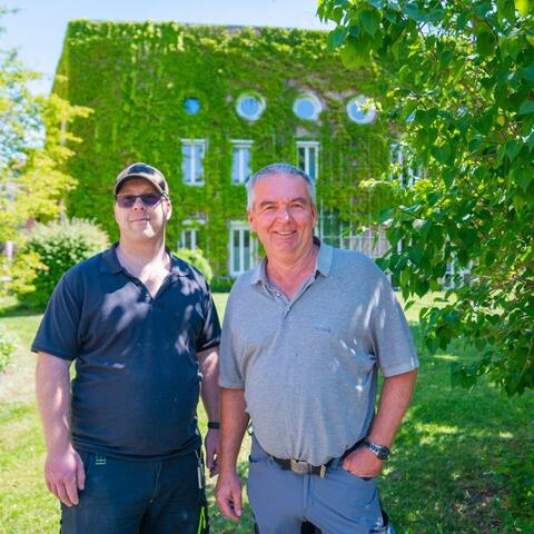 Zwei Männer im Freien – Zwei Mitarbeitende stehen lächelnd vor einem mit Efeu bewachsenen Gebäude