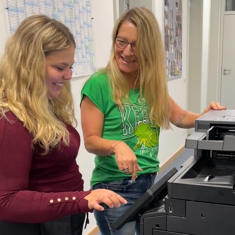 Kolleginnen am Kopierer – Zwei Mitarbeiterinnen bedienen gemeinsam einen Kopierer und lächeln