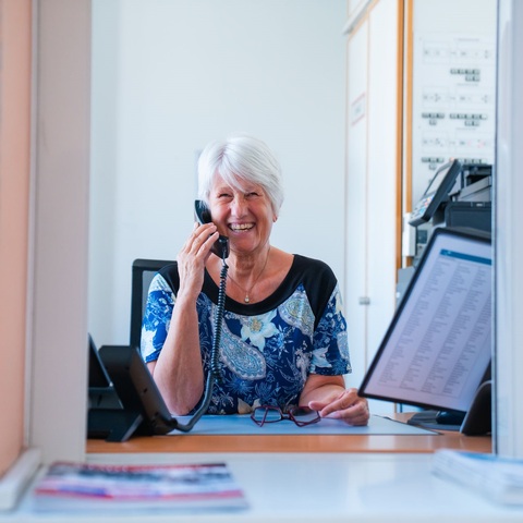 Ältere Dame mit Telefonhörer lächelt am Empfangsschalter