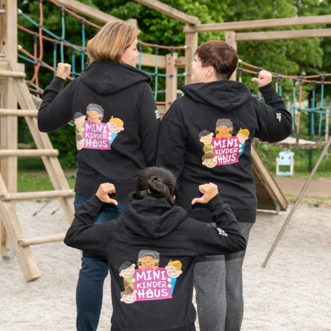 Drei Personen in schwarzen Kapuzenjacken mit dem Logo „Mini Kinder Haus“ auf dem Rücken posieren vor einem Klettergerüst auf einem Spielplatz.