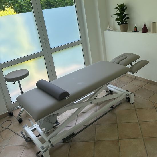 Behandlungsraum mit höhenverstellbarer grauer Therapieliege vor Fenster