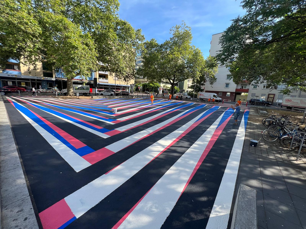 Künstlerische Bodenmarkierung auf einer großen Platzfläche mit breiten Streifen in Weiß, Blau, Pink und Schwarz, umgesetzt im öffentlichen Raum mit sichtbaren Markierungsarbeiten.