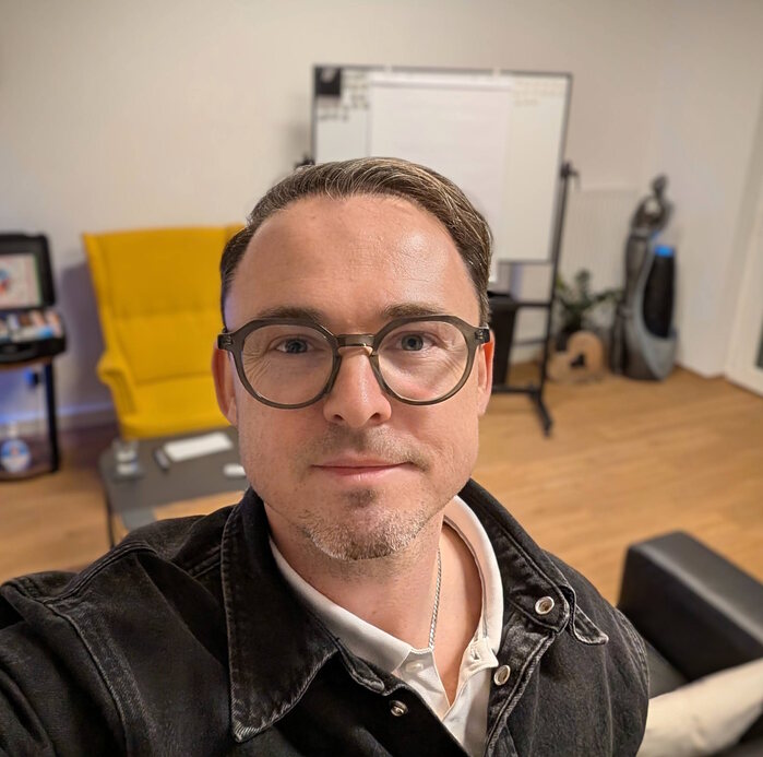 Mag. Streitberger sitzt auf einem auffälligen Ohrenstuhl. Er hält einen Stift und Moderationskarten in den Händen. Seine Körperhaltung ist aufrecht und aufmerksam. Er blickt in die Richtung der nicht sichtbaren Klient:innen und hört ihnen gespannt zu.