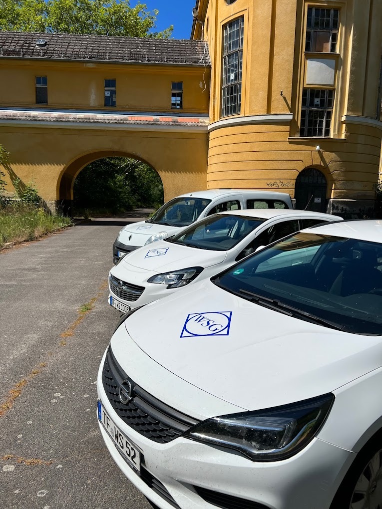 Mehrere weiße Einsatzfahrzeuge der WSG Zossen mit Firmenlogo auf einem Parkplatz vor einem gelben Gebäude.