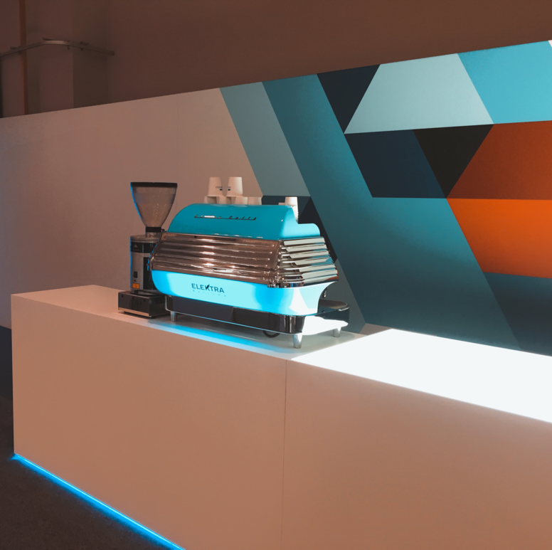 Zu sehen ist eine blaue Elektra Barlume Siebträgermaschine mit zwei Brühgruppen. Das Gerät hat die charakteristisch runde Retro‑Form der Barlume‑Serie und leuchtet in einem kräftigen, glänzenden Blauton. Die Maschine steht auf einer weißen Theke mit LED‑Beleuchtung, die das Gerät und die Arbeitsfläche hell ausleuchtet. Die Kombination aus blauem Metallgehäuse und weißer LED‑Theke erzeugt einen modernen, hochwertigen Eindruck, wie er typischerweise auf Messen oder Events eingesetzt wird.