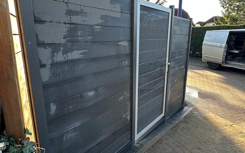 Sichtschutzzaun aus waagerecht verlaufenden, dunklen Holzpaneelen mit integriertem Eingangstor, montiert auf einem gepflasterten Bereich.