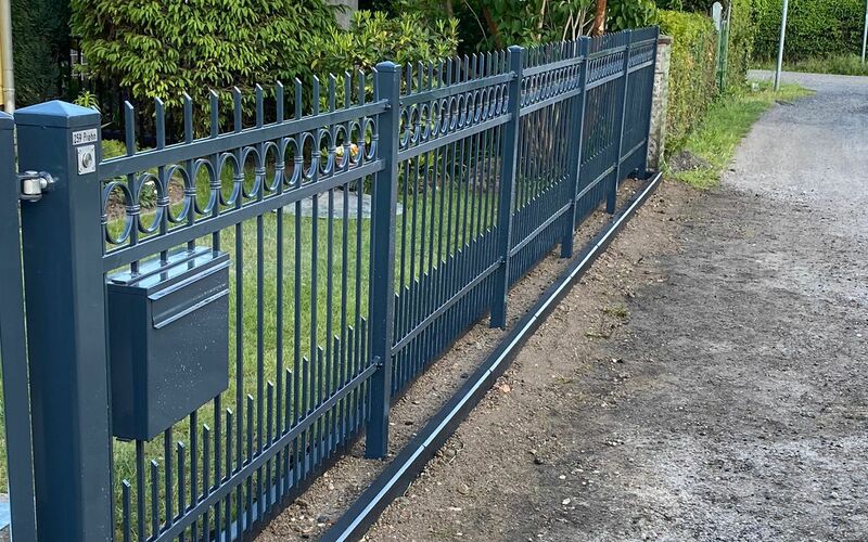 Dekorativer Metallzaun in Anthrazit mit Rundornamenten im oberen Bereich, integriertem Briefkasten, aufgestellt entlang eines gepflegten Grundstücks an einer Nebenstraße.