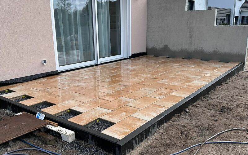 Halbfertige Terrasse mit beigefarbenen Natursteinplatten in regelmäßigem Muster, teilweise verlegt, mit schwarzer Randverkleidung; angrenzend an ein modernes Wohnhaus mit großen Terrassentüren.