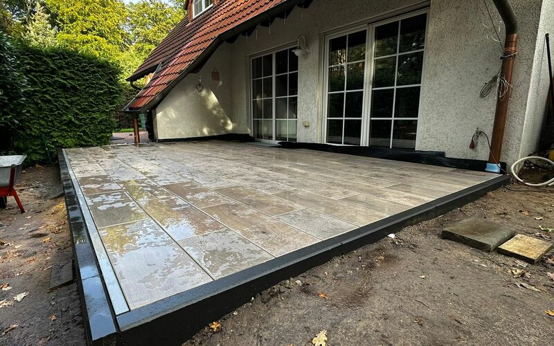 Fertiggestellte Terrasse mit großformatigen Feinsteinzeugplatten in Natursteinoptik und dunkler Rahmenverkleidung, gelegen vor einem Wohnhaus mit Sprossenfenstern und einer Hecke als Gartenbegrenzung.