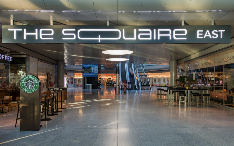 Eingang zur Einkaufspassage „The Squaire East“ mit Starbucks und Rolltreppe
