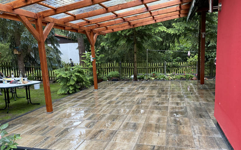 Große Terrasse mit strukturierten Feinsteinzeugplatten in Holzoptik/Granit, überdacht mit einer Pergola aus Holz, angrenzend an ein rotes Wohngebäude und einen Naturgarten mit Bäumen und Holzzaun.