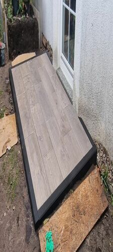 Kleine, moderne Terrasse mit hellgrauen Feinsteinzeugplatten in Holzoptik, eingefasst von einem schmalen, dunklen Rahmen, direkt an eine Hauswand angrenzend, der Boden rundherum ist noch unbearbeitet.