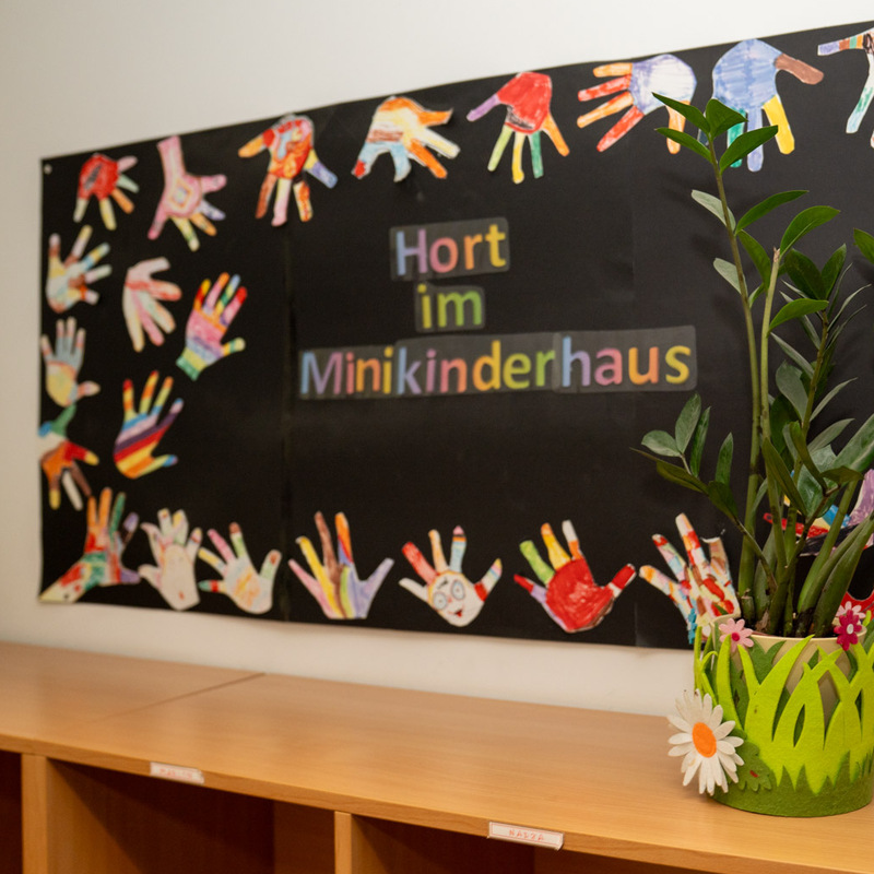 Schwarzes Poster „Hort im Minikinderhaus“ mit bunten Handabdrücken an einer Wand