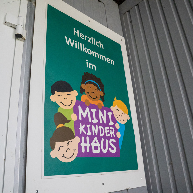 Eingangsschild „Herzlich Willkommen im Minikinderhaus“ mit lachenden Kinderillustrationen