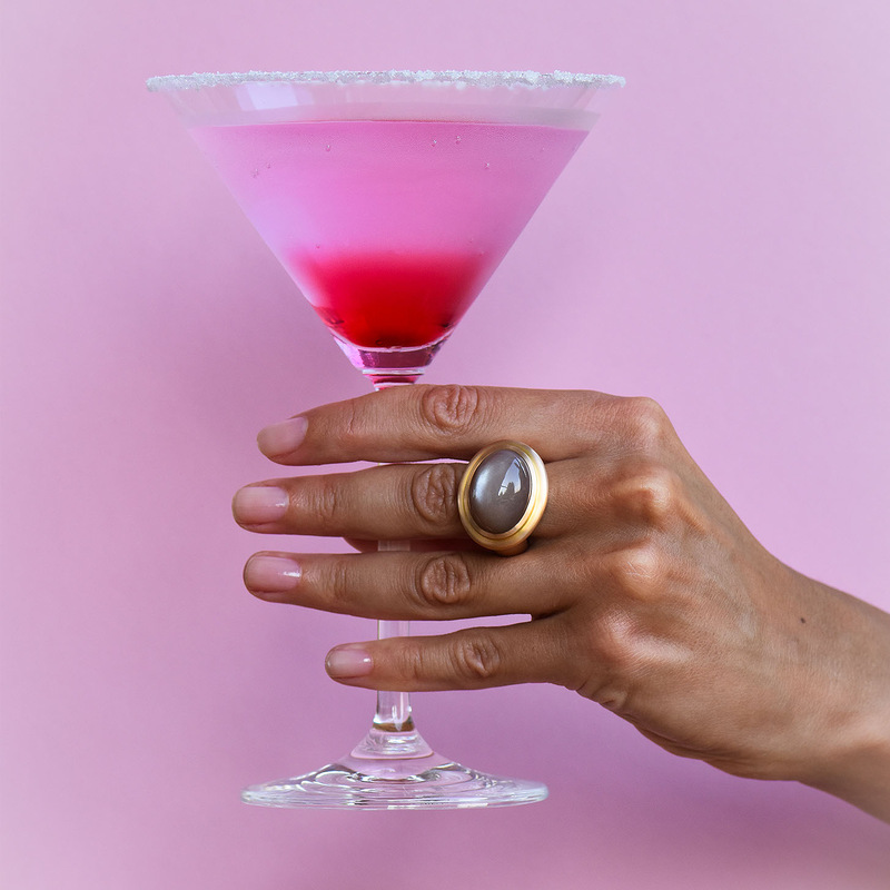 Hand mit auffälligem Ring und Cocktailglas vor rosafarbenem Hintergrund