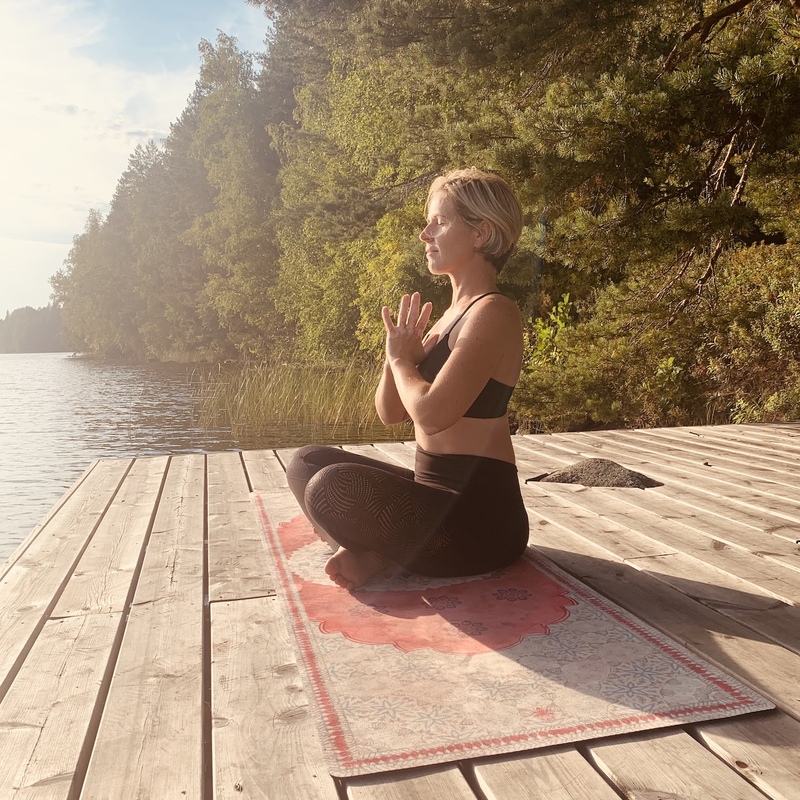 Frau in Meditationshaltung im Schneidersitz auf Steg vor einem See im Sonnenlicht