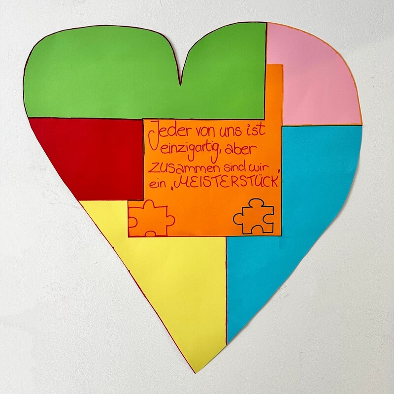 Buntes Papierherz mit Puzzleteilen und der Aufschrift „Jeder von uns ist einzigartig, aber zusammen sind wir ein Meisterstück“