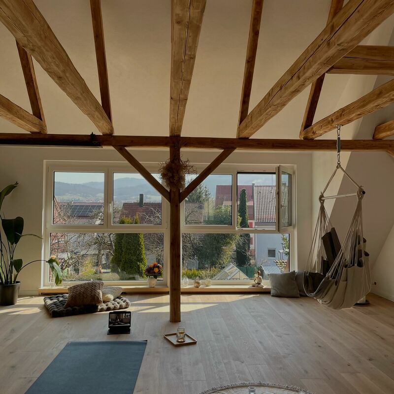 Lichterfüllter Yoga-Raum mit Blick durch große Fenster auf bewaldete Hügel