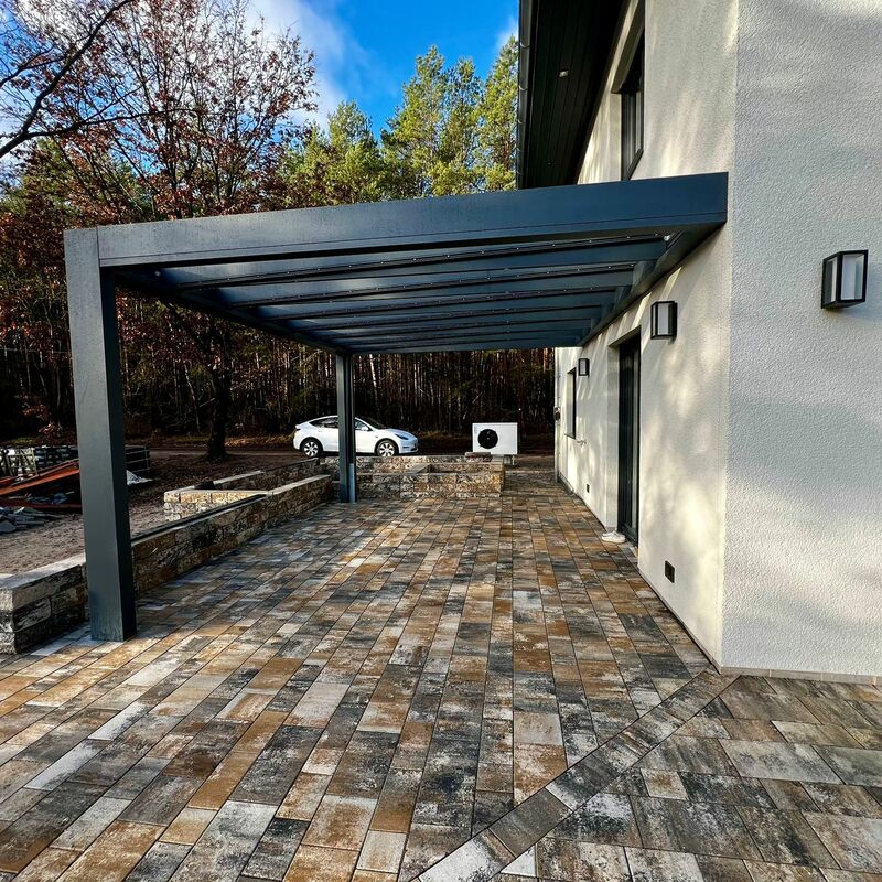 Große Terrasse mit gemusterten Pflastersteinen in Braun- und Grautönen, überdacht von einer modernen, offenen Pergola aus dunkelgrauem Metall, angrenzend an ein helles Wohnhaus.
