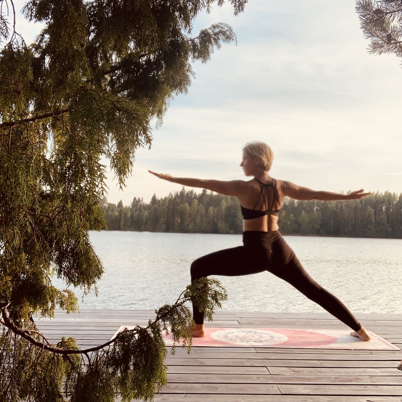 Frau in Krieger-II-Yoga-Position auf Steg vor Waldsee unter bewölktem Himmel