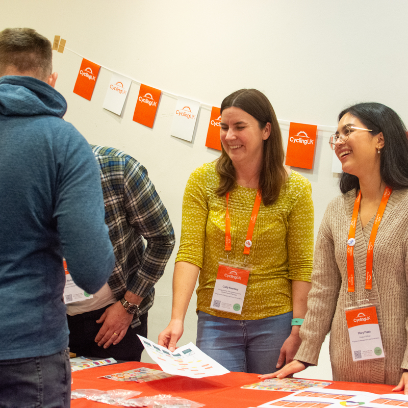 Staff sharing their recent work for an internal exhibition style networking event

(Picture of Cally and Mary standing at a table smiling, wearing orange Cycling UK lanyards, and talking to a staff member who faces them)
