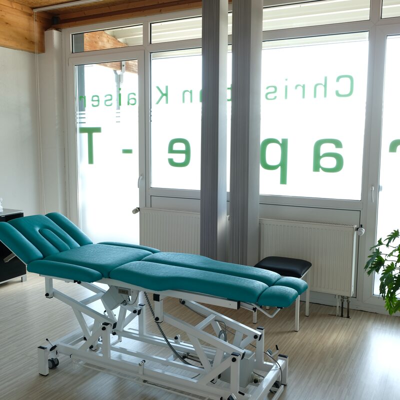 Behandlungsraum mit grüner Therapieliege vor großer Fensterfront