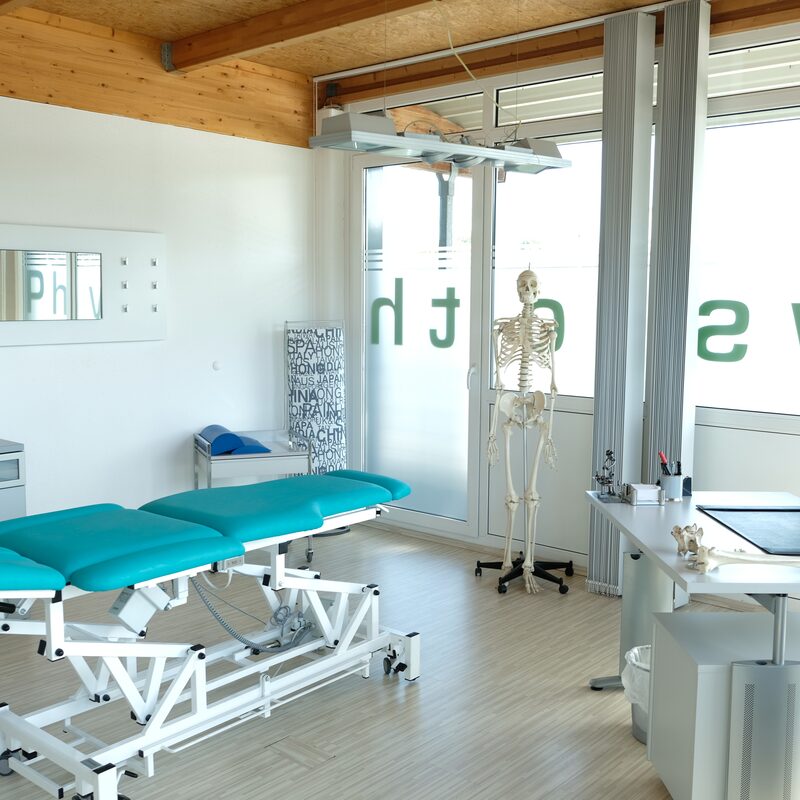 Heller Behandlungsraum mit Therapieliege, Schreibtisch und Skelettmodell