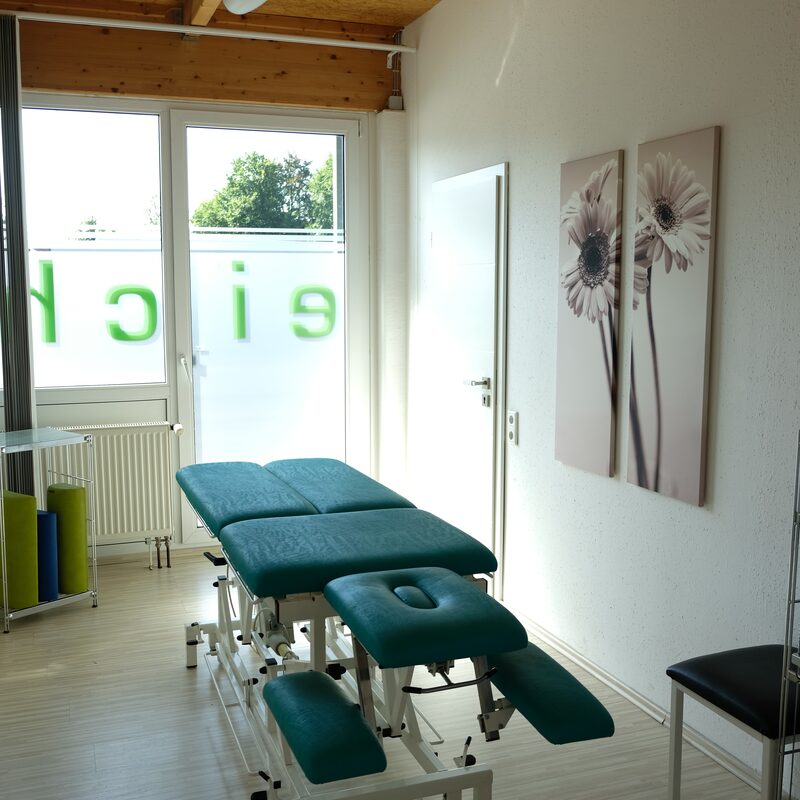 Behandlungsraum mit grüner Therapieliege und Blumenbildern an der Wand