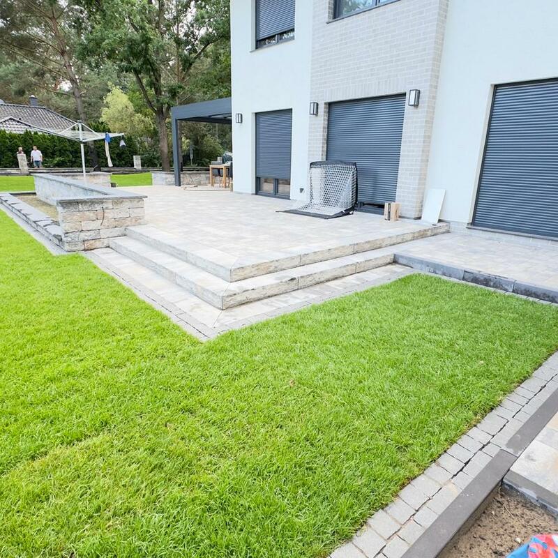 Terrasse mit hellen Natursteinplatten und breiten Treppenstufen, angrenzend an eine gepflegte Rasenfläche, an einem modernen Wohnhaus mit weißen Wänden und geschlossenen Rollläden.