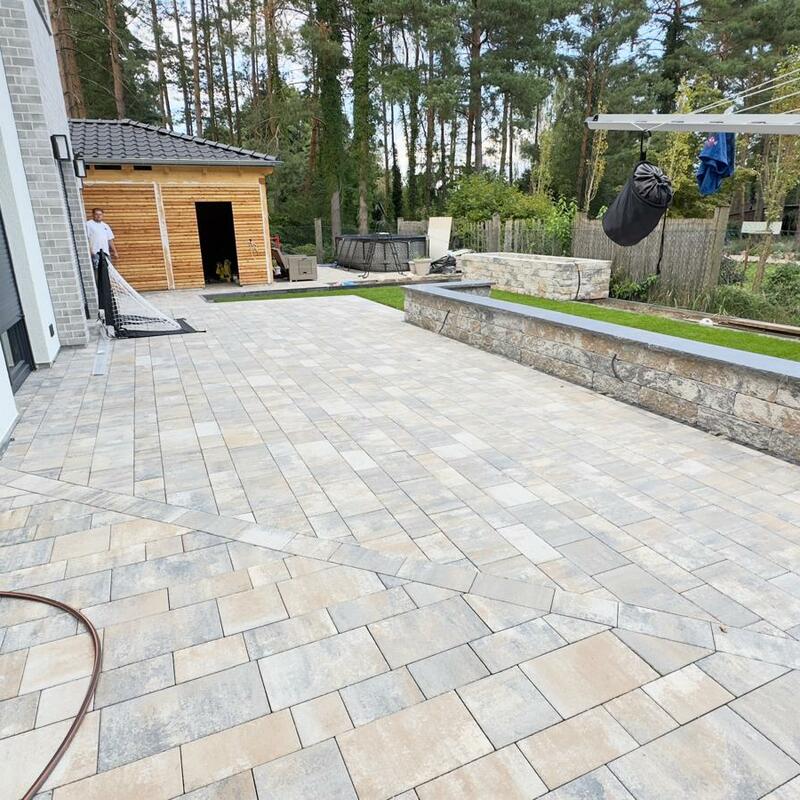 Weitläufige Terrasse aus hellen Pflastersteinen in Naturtönen, ergänzt durch eine niedrige Natursteinmauer mit dunkler Abdeckplatte, angrenzend an einen Garten mit Holzhaus und umgeben von Bäumen.