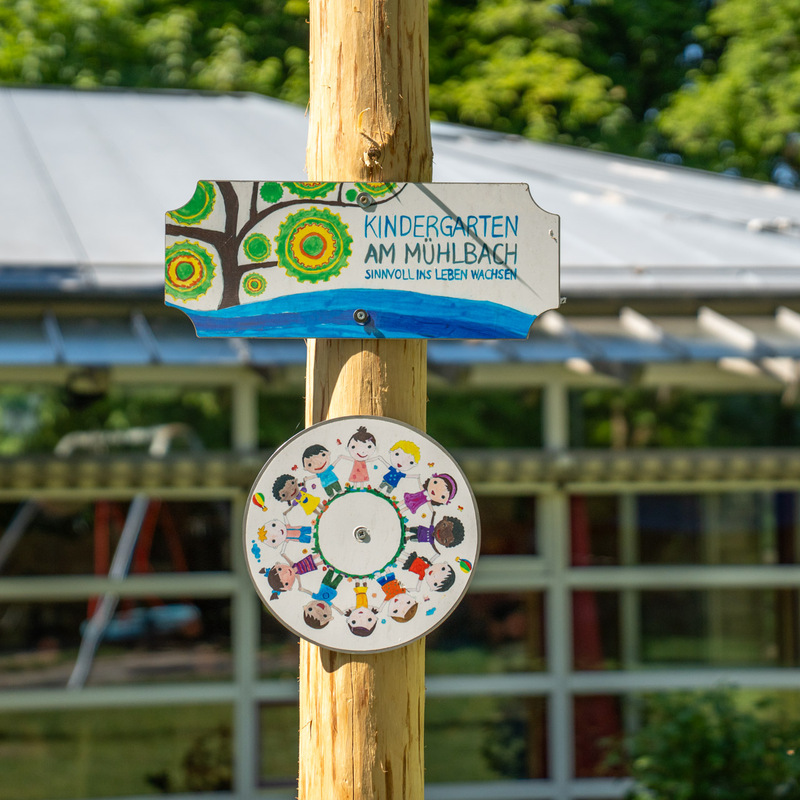 Kindergarten-Schild – Holzschild mit der Aufschrift „Kindergarten am Mühlbach“ und gemalten Kinderfiguren