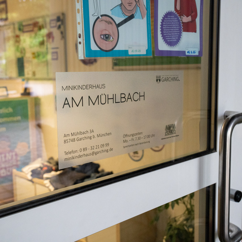 Eingangsschild Minikinderhaus – Glastür mit Beschriftung „Minikinderhaus Am Mühlbach“ und Kontaktdaten