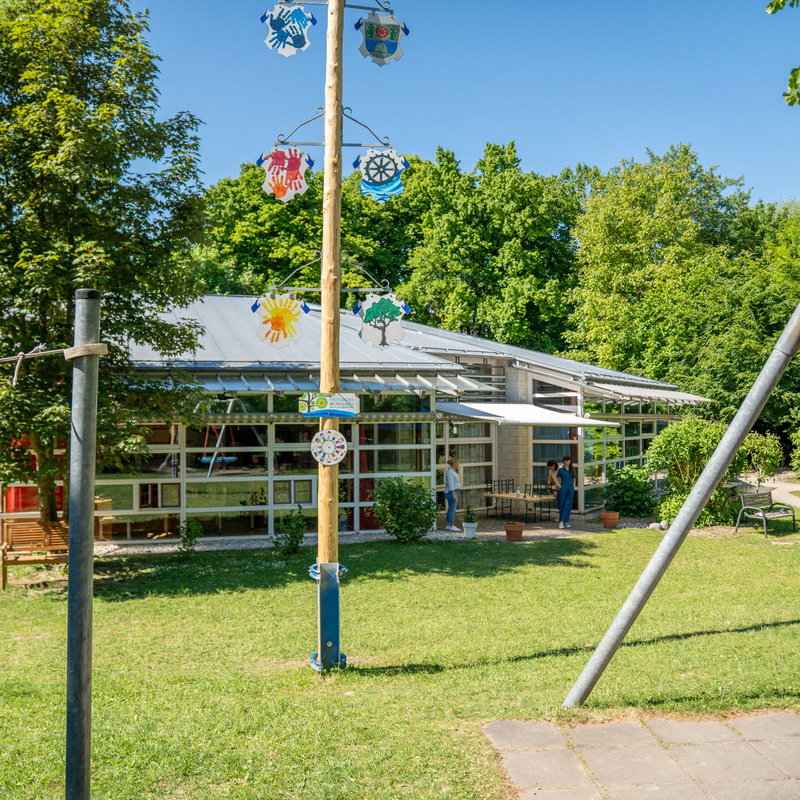 Kindergarten im Grünen – Außenansicht eines Kindergartens mit Spielplatz und bunt bemaltem Maibaum