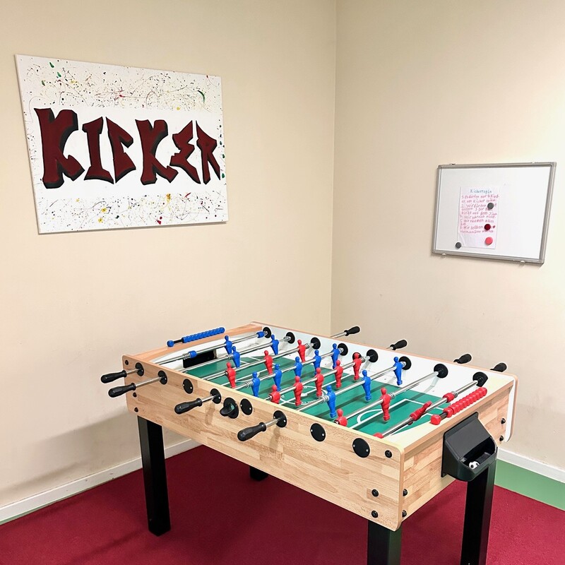 Kickertisch auf rotem Teppich mit Wandbild „Kicker“ im Hintergrund