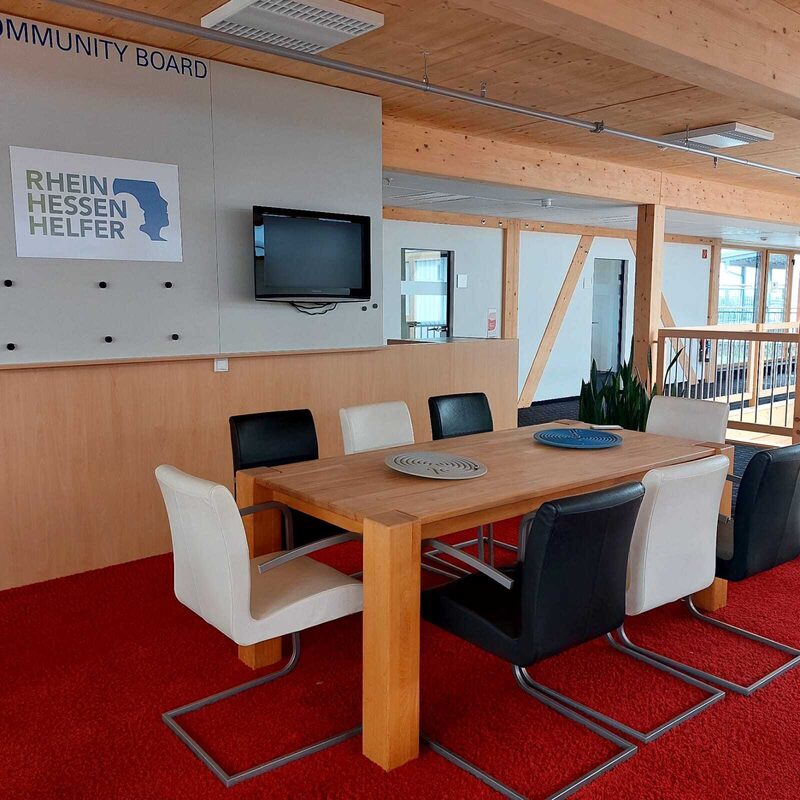 Tisch mit mehreren Stühlen vor einer Wand mit Fernseher und Logo „Rheinhessen Helfer“