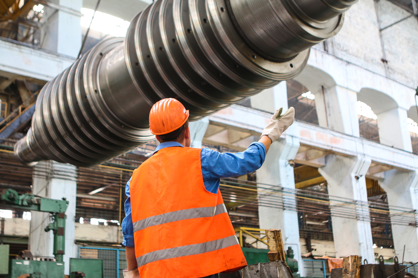 Mitarbeiter mit Schutzhelm und Warnweste prüft eine große Maschinenwelle in einer Industriehalle